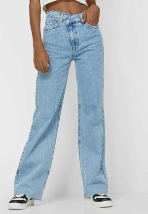 Vrouw draagt lichtblauwe, hooggetailleerde jeans met wijde pijpen en zwart-witte sneakers, met een hand in de zak tegen een effen achtergrond.