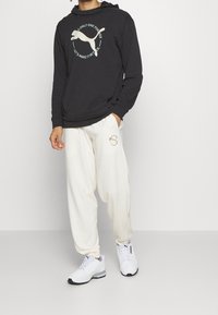 Man som bär en svart hoodie och vita sweatpants med Puma-logotyp, står med en hand i fickan och bär vita Puma-sneakers.