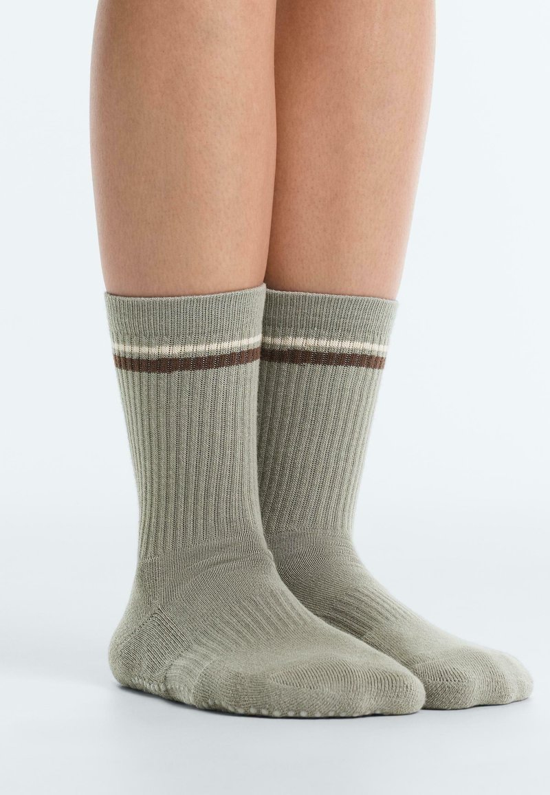 Noge, obute v svetlo zelene nogavice s srednjo riso, z belimi in rjavimi črtami blizu vrha, stojijo na beli površini.