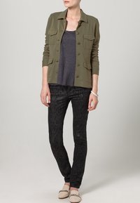 Olive green button-up jacket over a dark gray top, paired with black patterned skinny pants and light beige slip-on shoes.