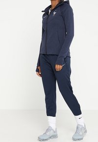 Marinblå sportjacka med dragkedja, huva och Frankrikes emblem; kombinerad med marinblå byxor och ljusgrå sneakers med vita detaljer och svart logotyp.