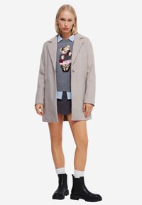 Lichtgrijze wollen blend jas met notch-kraag, gelaagd over een grijze sweater met een berenontwerp, gecombineerd met een blauwe blouse en zwarte enkellaarzen.