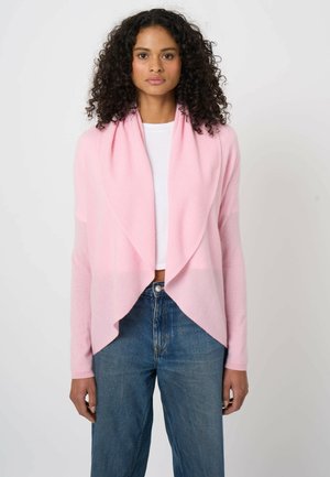 Kobieta o kręconych włosach, ubrana w jasnoróżowy sweter z otwartym przodem, biały top i niebieskie dżinsy, stojąca na tle jednolitego białego tła.