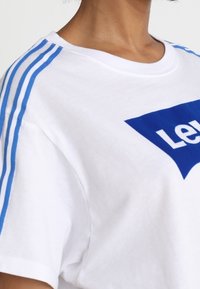 Camiseta de algodón blanca con acentos de rayas azules en las mangas y un gran logo gráfico azul en la parte delantera. Textura suave con un corte relajado.