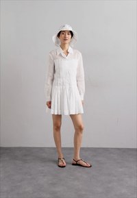 Abito bianco plissettato con scollo a collare, abbinato a un cappello a secchiello bianco in crochet e sandali piani neri. Tessuto morbido e design minimalista.