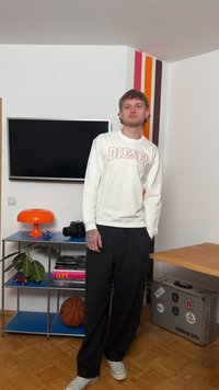 Giovane uomo con felpa bianca Diesel e pantaloni neri in piedi in una stanza con una TV, una lampada arancione, un pallone da basket e una valigia argentata con adesivi.