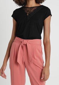 Top negro de manga corta con escote en V de encaje combinado con pantalones de talle alto en rosa empolvado que cuentan con un detalle de cinturón anudado.