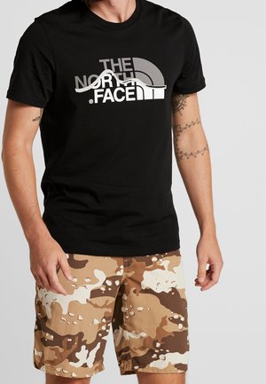 Mann trägt ein schwarzes The North Face T-Shirt und beige-braun-weiß getarnte Shorts, steht vor einem einfarbigen Hintergrund.