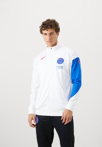 Hvid zip-up sportjakke med blå ærmer, forsynet med et rundt logo og teksten "QATAR". Lavet af glat, letvægtsmateriale. Sorte bukser.