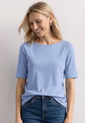 Mujer rubia sonriendo y mirando hacia abajo, vistiendo una camiseta de manga corta azul claro y vaqueros azul oscuro sobre un fondo liso.