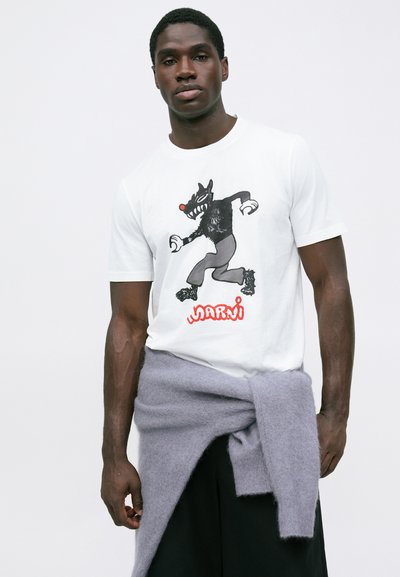 Balta kokvilnas T-krekls ar melnu izplūduša stila vilka zīmējumu ar sarkanu degunu, kopā ar gaiši pelēku džemperi, kas sasiets ap jostasvietu.