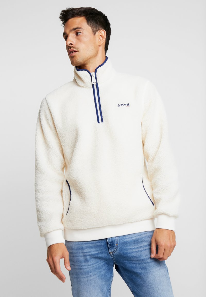 Mand iført en cremefarvet fleecepullover med marineblå kant og lynlås, parret med blå jeans, ser mod venstre mod en ensfarvet baggrund.
