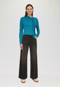 Türkisfarbene langärmlige Bluse mit einem gebundenen Detail am Ausschnitt, kombiniert mit hochgeschnittenen schwarzen weiten Jeans und beigen Plateauschuhen.