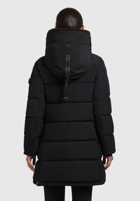 Melnā pūkainā jaka ar lielu kapuci, ribotām detaļām un teksturētu apkakli. Iekļauj logo pievienotu siksnu aizmugurē un pāršūtas daļas.