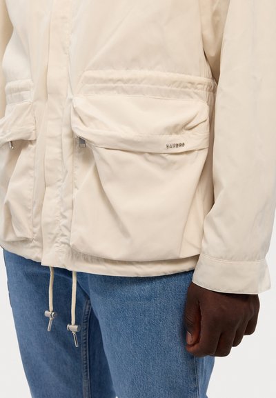 Svetlo bež jakna z velikimi sprednjimi žepi, z vrvico in logotipom Sandro, oblečena z modrimi zanosom in temno kožnim dlanom vidnim ob strani.