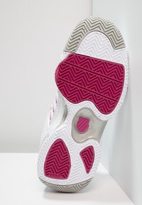 Sålen på en vit sportskor med rosa, grå och vita sicksack-mönster och en liten sköldlogotyp nära fotvalvet.