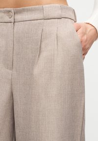 Κοντινό πλάνο σε εκρού ραμμένο παντελόνι με κουμπί και πιέτες, εμφανίζοντας ένα χέρι να ακουμπά μέσα στην δεξιά τσέπη.