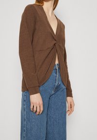 Maglione marrone a maniche lunghe con scollatura a V profonda e dettaglio ancorato sulla parte anteriore. Tessuto morbido, maniche aderenti e orlo drappeggiato. Abbinato a jeans a vita alta.