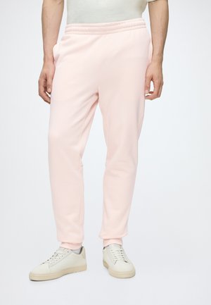 Personne portant un pantalon de jogging rose clair et des baskets blanc cassé, debout sur fond blanc.