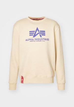 Beige sweatshirt featuring a purple "Alpha Industries" logo, crew neck, ribbed cuffs, and a red tag at the hem. Soft fabric texture.