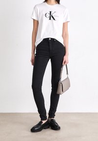 Camiseta blanca de algodón con el logo "CK" en negro, jeans de tiro alto negros y mocasines negros. Sosteniendo un pequeño bolso gris con una correa.