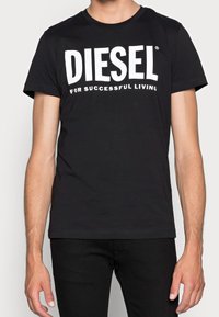 Muž v černém tričku Diesel s bílým textem "DIESEL for Successful Living", spojený s černými kalhotami, stojí proti jednoduchému pozadí.
