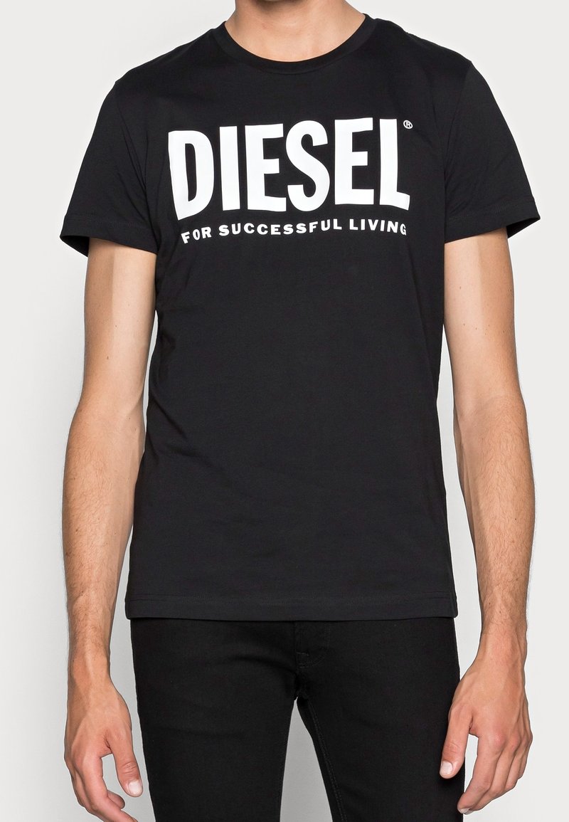 Muž v černém tričku Diesel s bílým textem "DIESEL for Successful Living", spojený s černými kalhotami, stojí proti jednoduchému pozadí.