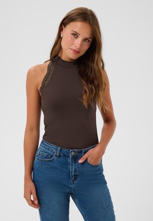 Giovane donna con capelli lunghi che indossa un top senza maniche marrone scuro con collo alto e jeans blu, con una mano in tasca, in piedi davanti a uno sfondo semplice.