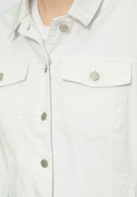 Witte denim jas met metalen drukknoopsluitingen, twee borstzakken en een gestructureerde kraag. Soepele textuur met subtiele stikdetails.