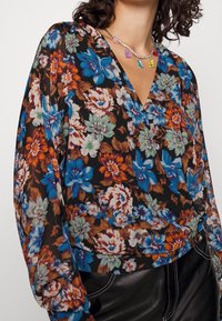 Blouse florale avec un fond noir orné de fleurs multicolores en bleu, orange et vert. Manches longues et col en V.