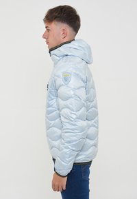 Giacca imbottita azzurra chiara con finitura lucida, cappuccio e patch logo con accenti gialli sulla spalla. Presenta un design trapuntato curvo.