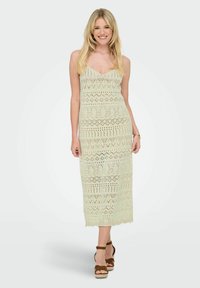Rochie maxi din croșetat de culoare verde deschis, cu decolteu în V, modele complexe și o siluetă ajustată. Asortată cu sandale cu brâu la gleznă de culoare maronie.