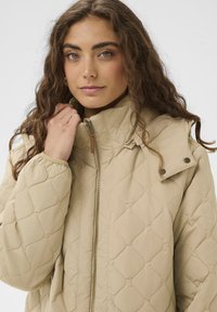 Chaqueta acolchada beige con cuello alto, cuenta con cierre de cremallera y botones de presión. Textura suave con un distintivo patrón de diamantes.