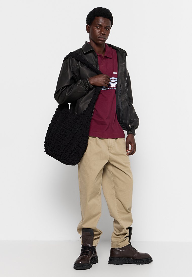 Czarna skórzana kurtka bomberka na maroonowej koszulce polo, khaki spodniach i brązowych butach wojskowych. Niesie teksturowaną czarną torbę.