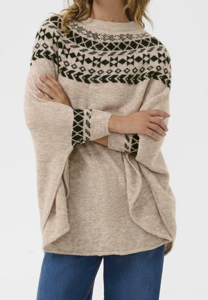 Frau trägt einen beigen Strickpullover mit dunklen geometrischen Mustern am Yoke und an den Bündchen, die Arme verschränkt, kombiniert mit blauen Jeans.