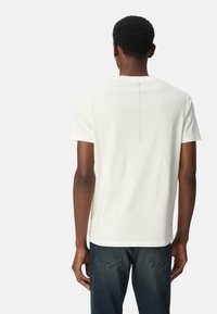 Camiseta blanca de algodón con cuello redondo, mangas cortas y un detalle de costura vertical en la parte posterior. Llevada con jeans de mezclilla oscura.