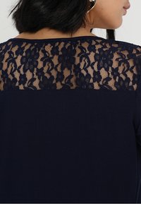 Marinblus med en blommig spetsbakpanel med intrikata mönster. Framsidan är hel och slät, vilket kontrasterar med den texturerade spetshyden.