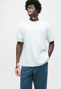 Ανοιχτό μπλε φαρδιά μπλούζα με ρολαρισμένα μανίκια, διακοσμημένη με διακριτικό λογότυπο στο στήθος. Συνδυάζεται με σκουρόχρωμο παντελόνι jogger και υφασμάτινο φόντο.