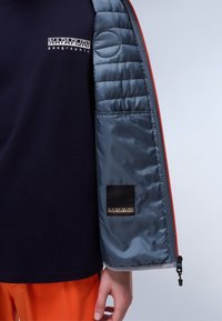 Navy jacket with a gray quilted lining, orange zipper, and black logo patch on the sleeve. Smooth texture with insulated padding.