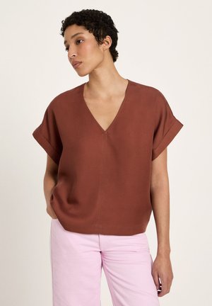 Woman with short curly hair wearing a rust-colored V-neck top and light pink pants, standing against a plain white background.