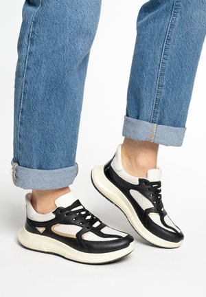 Feet wearing black and white sneakers with thick soles and rolled-up blue jeans against a white background.