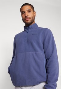 Pullover in pile con mezza zip di colore blu scuro con finitura testurizzata, dotato di un colletto alto e un discreto logo sul petto.