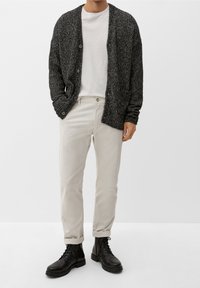 Cardigan nero a maglia sopra una maglietta grigia chiara, abbinato a pantaloni beige arrotolati e stivali neri con lacci. Design semplice e strutturato.