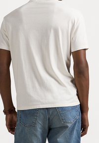 Beżowa bawełniana koszulka z luźnym krojem, krótkimi rękawami i prostym, nieozdobionym tyłem, w połączeniu z niebieskimi dżinsami.