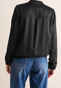 Veste bomber noire avec une texture lisse, ourlet et poignets élastiques. Associée à un jean en denim bleu avec des coutures visibles et des poches.