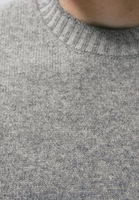 Grijze gebreide trui met een geribde halslijn, zachte textuur en een gespikkeld patroon. Close-up weergave die de details van de stof benadrukt.