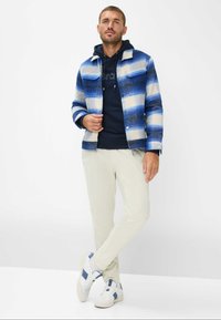Giacca in flanella a quadretti blu e bianchi, felpa blu navy sotto, pantaloni in velluto a coste color beige chiaro e sneakers bianche con accenti blu.