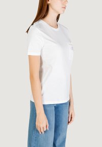 Camiseta de algodón blanca con mangas cortas, cuello redondo y un bolsillo frontal que presenta un colorido diseño bordado. Combinada con unos jeans de color azul claro.