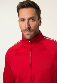 Giacca zip-up rossa con colletto alto, logo ricamato sul petto e strisce bianche lungo le spalle. Tessuto dalla texture liscia.