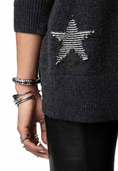Pull gris foncé avec un tricot texturé, présentant un design d'étoile rayée blanche dans le dos. Des bracelets métalliques ornent le poignet, avec un pantalon noir.